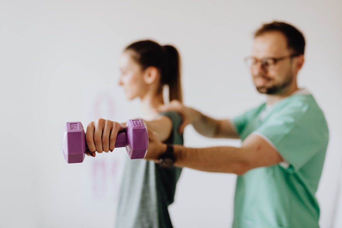 A male physical therapist assistant with glasses and a beard helping his patient lift a small pink weight.  Physical Therapist Assistant Associate Degrees