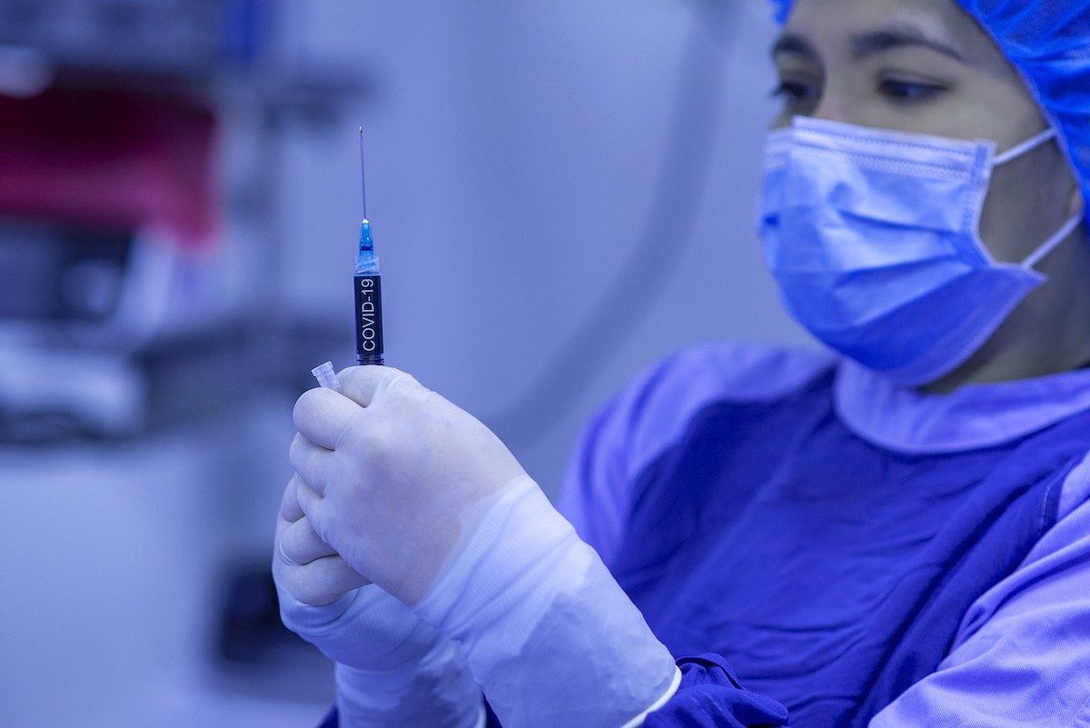 A nurse wearing blue hospital overalls and face mask while holding a Covid-19 vaccine How To Get Into Nursing School