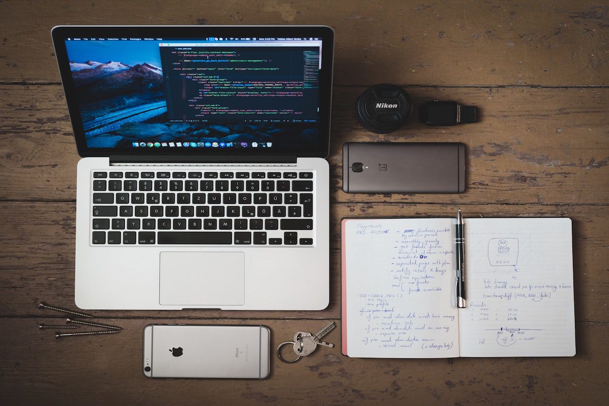 An image of a silver MacBook with colorful code on it placed next to a pair of Nikon headphones, two cell phones, a notebook, a pen, and keys on a table. 