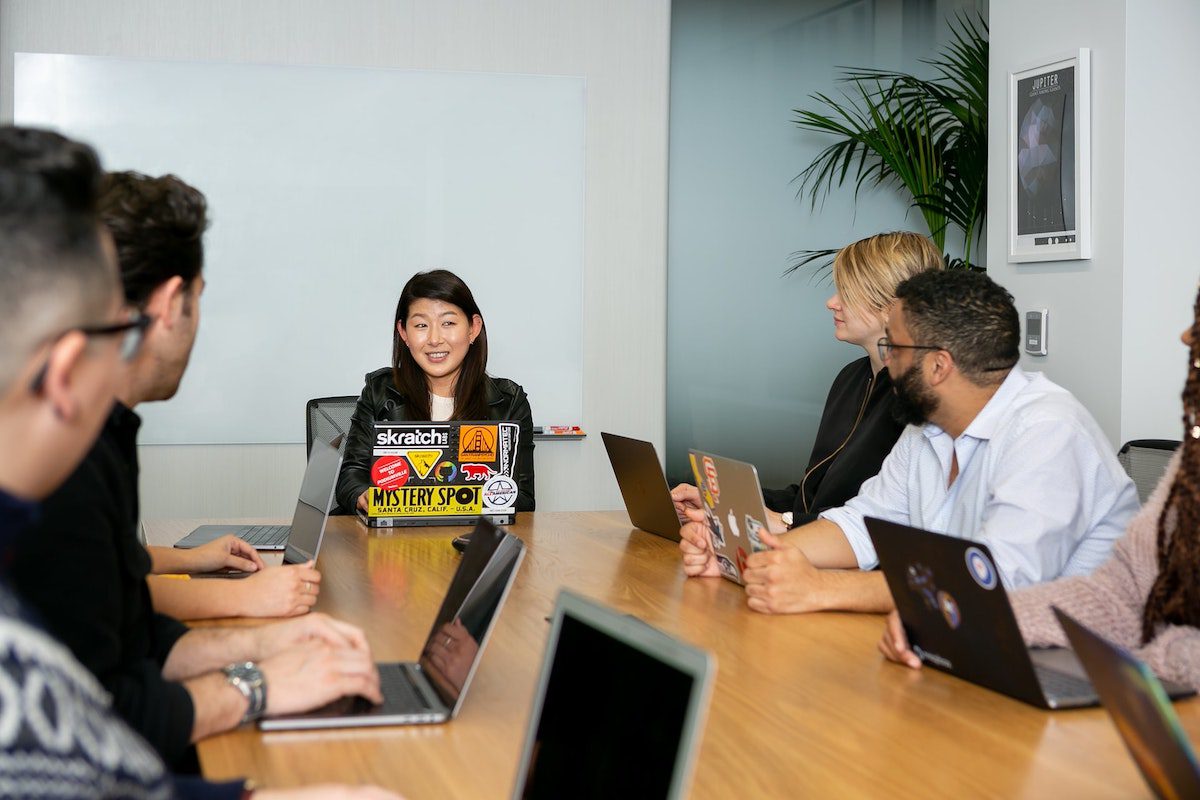 Six tech workers with laptops sitting around a meeting table, listening to someone speak