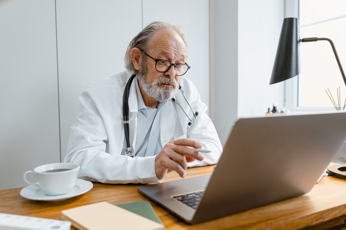 Alt-text: An older doctor wearing glasses sitting in front of a laptop computer with a large cup of coffee.