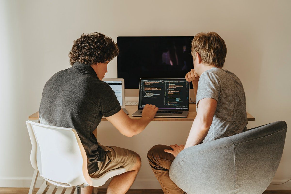 Two casually dressed men sitting in chairs at a desk looking at a laptop screen