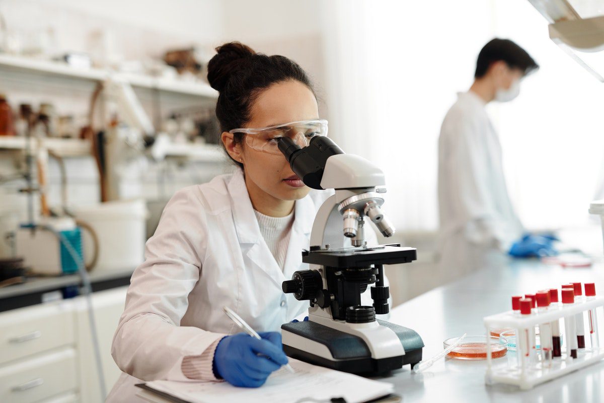 Female scientist in white lab coat using a microscope and writing notes.