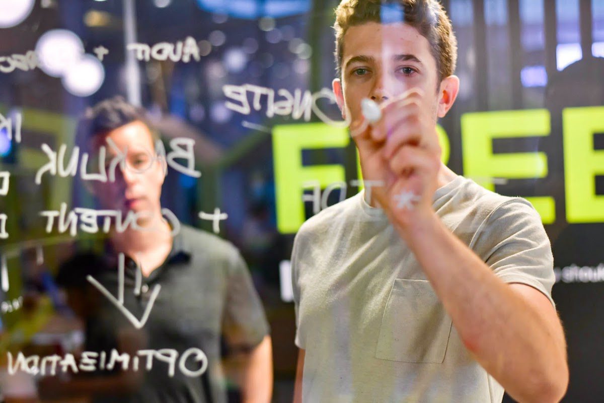 Two male business intelligence analysts brainstorming business strategies, with one of the men writing on glass with a white marker.