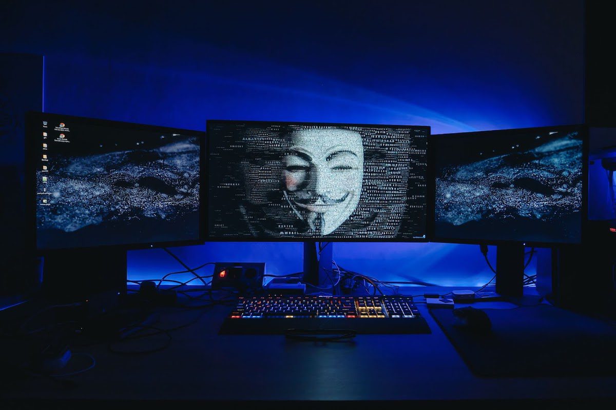 Three computer monitors in a dark room with an anonymous mask displayed on one screen.
