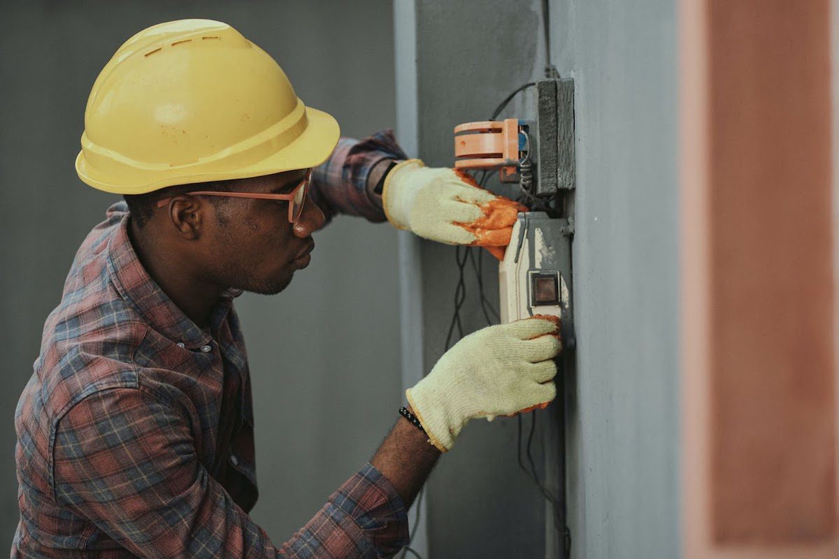 An electrician in a yellow hard hat and white gloves fixing a fuse box.