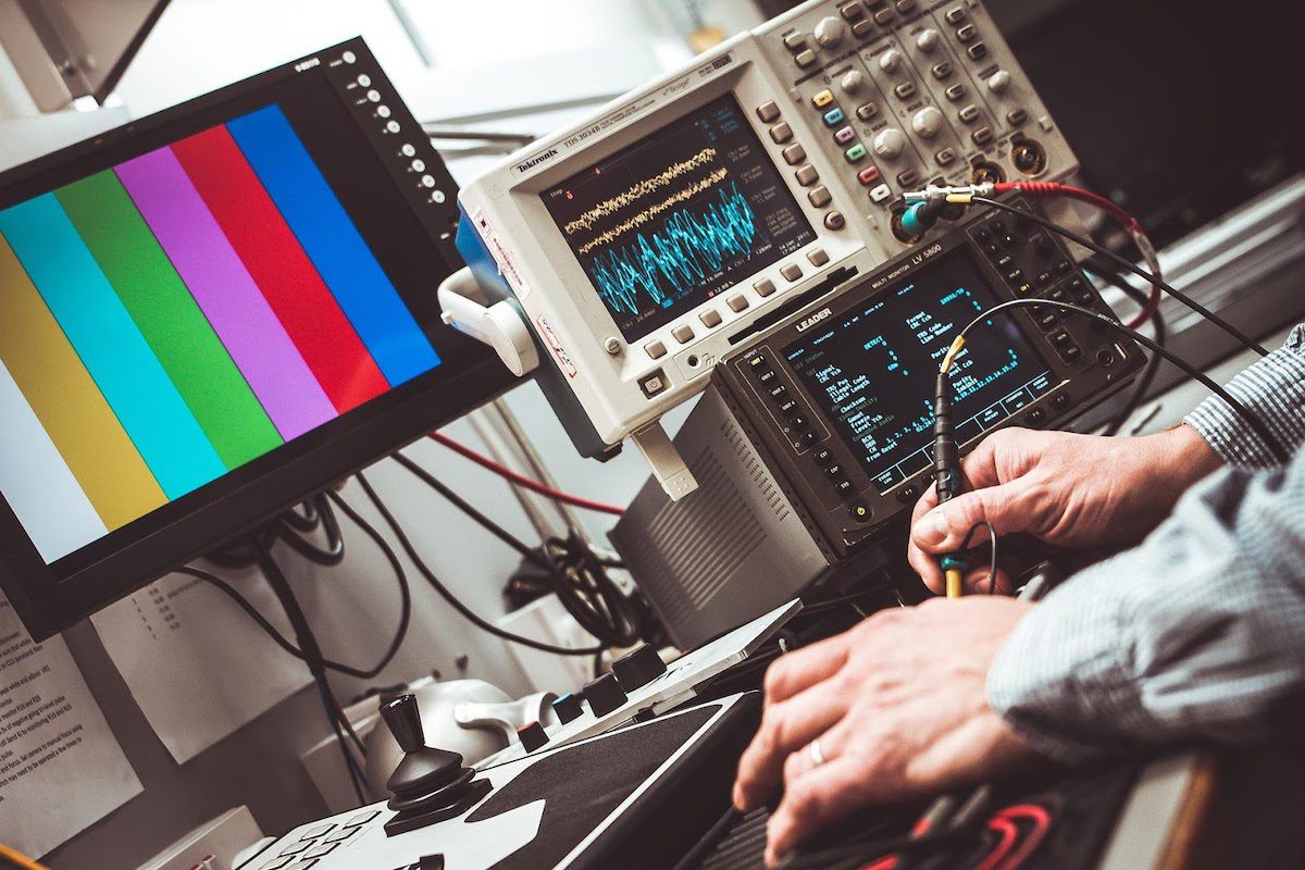 A pair of hands holding tools with colored lines on a monitor and electrical equipment