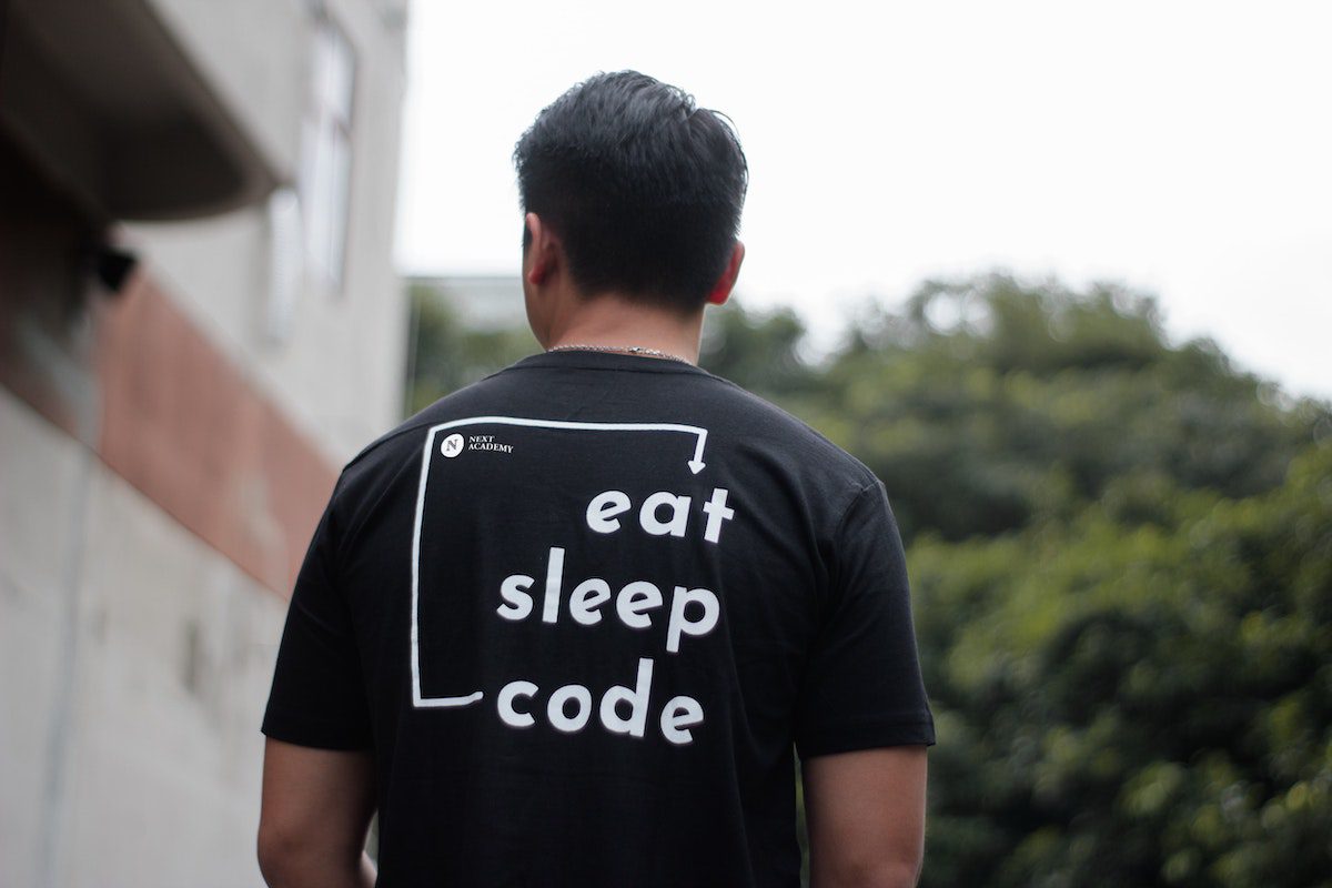 A man with his back to the camera wearing a black t-shirt that says "eat, sleep, code", enjoying his time at a paid coding bootcamp.