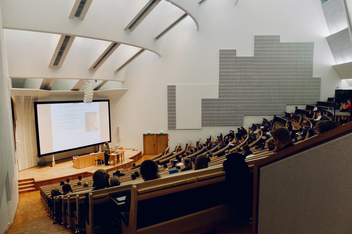 Students sit in a large lecture hall while a professor stands at the front, giving a presentation on a large screen.