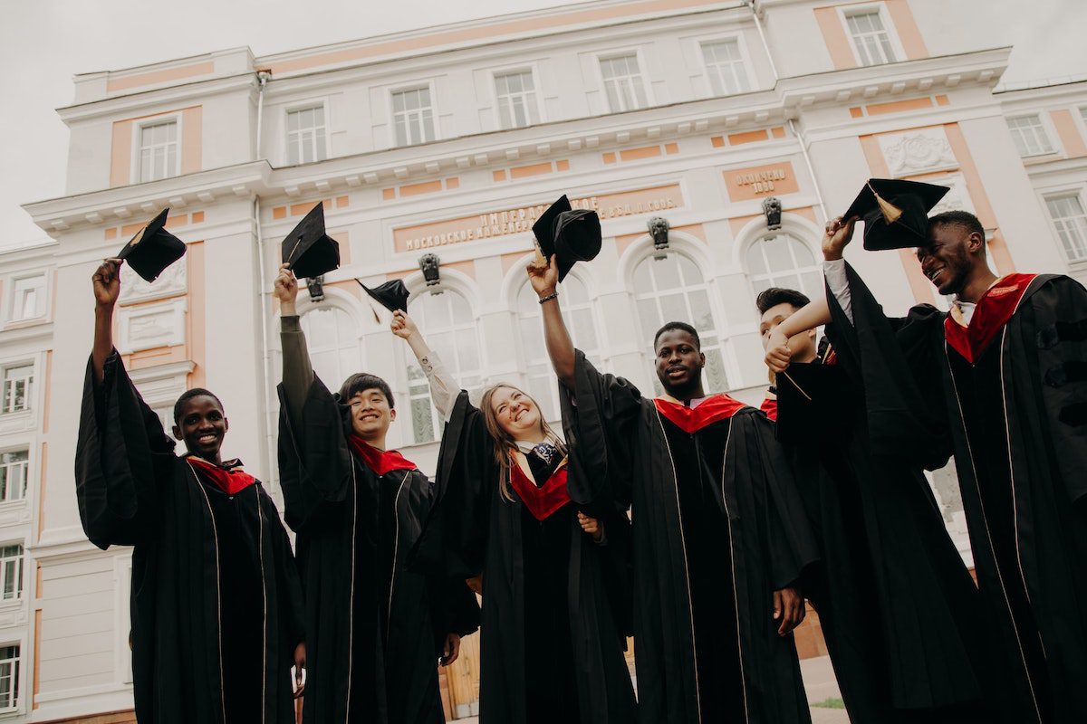 Six graduating college students stand in a line outside in front of a large building. They are wearing convocation regalia and holding their caps in the air.