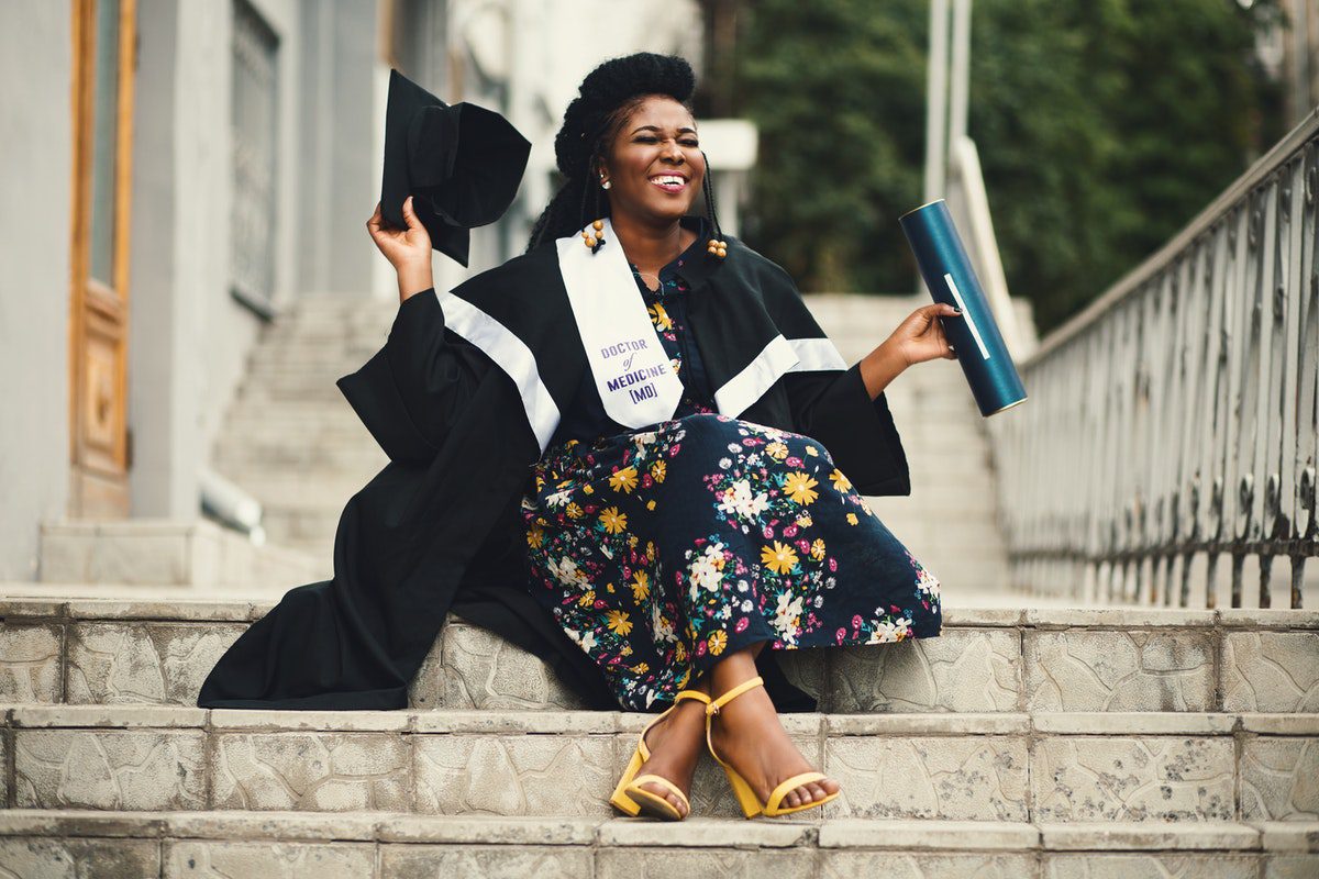 A graduate sitting on a staircase in graduation clothing smiling and holding a graduate cap and her diploma.