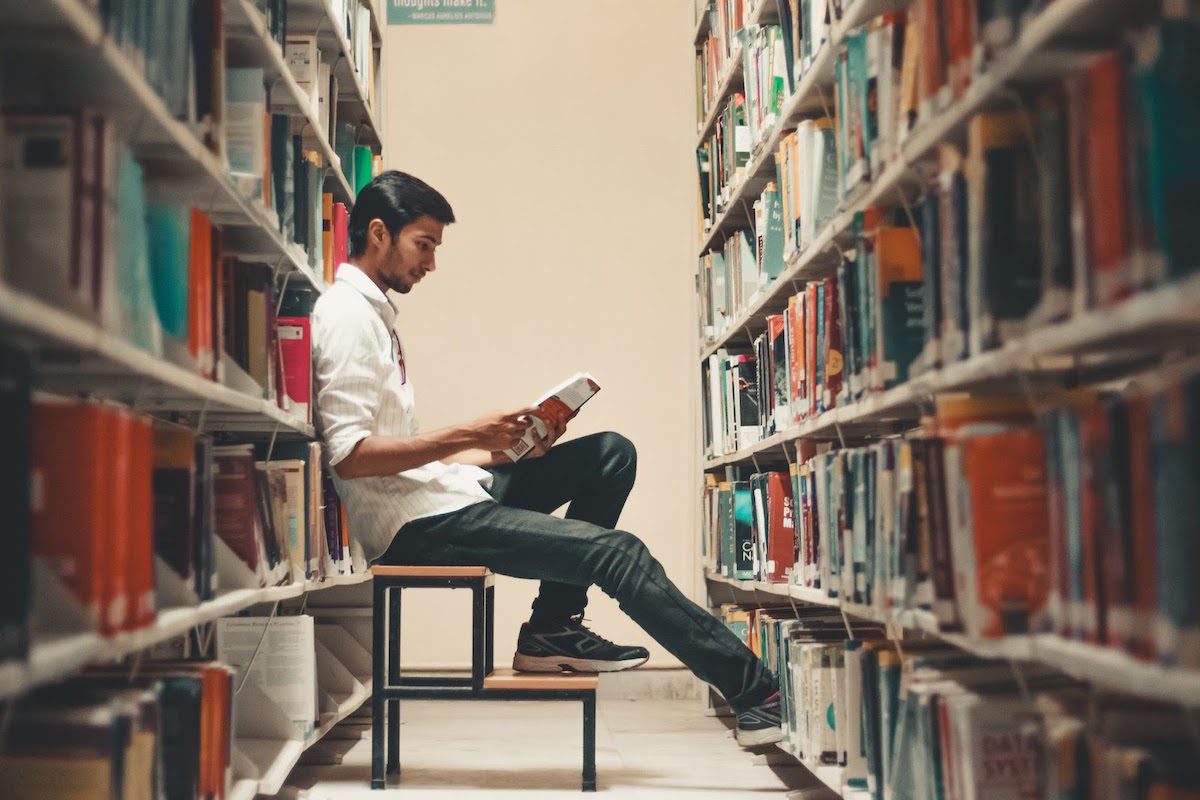 A young man sitting on a stool reading a book in a university library 