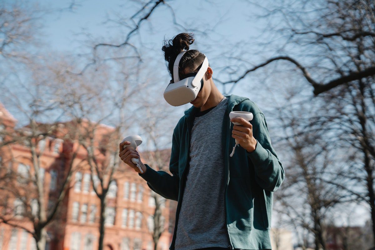 A man outside wearing virtual reality goggles and holding a controller in each hand.