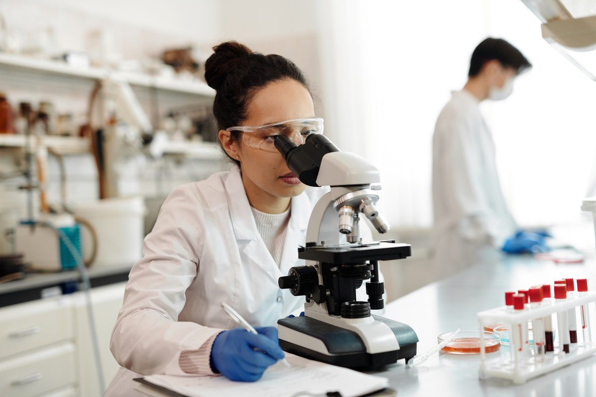 A scientist is white lab coat using a microscope and taking notesBiologists usually work in a lab where they conduct their research.