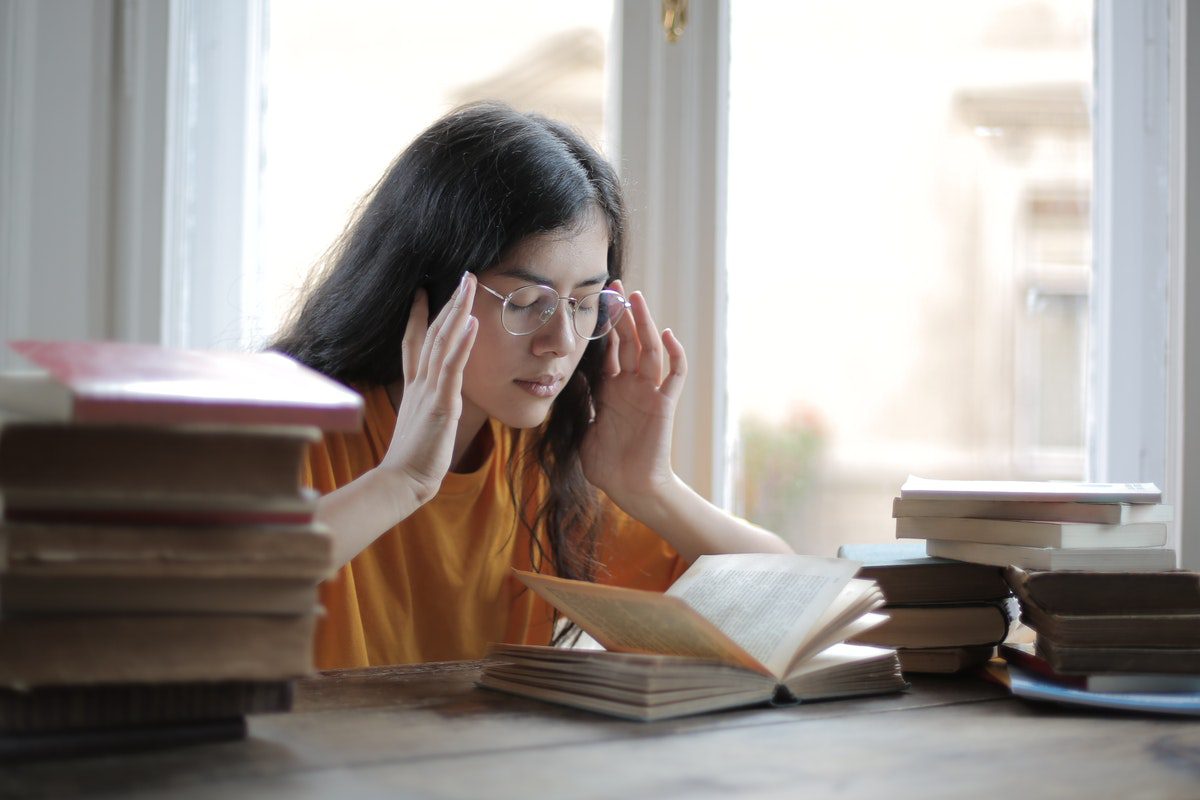 Student rubbing their temples with eyes closed, at a desk surrounded by stacks of books.