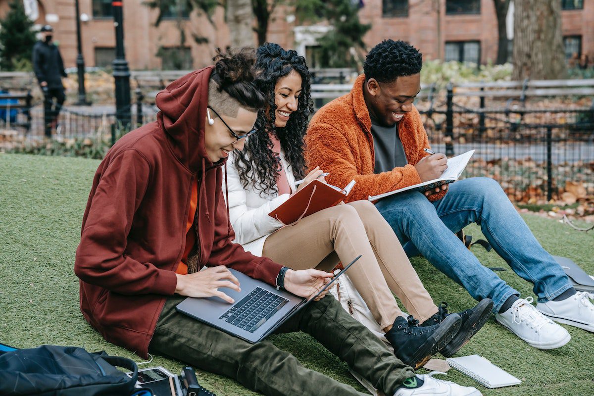 Three students sit on the grass and write their assignments