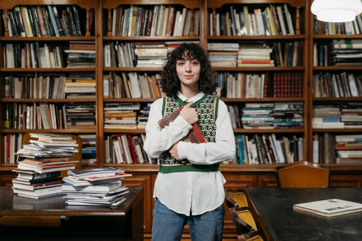 An introvert wearing a green vest and a white dress shirt while holding a book in a library.