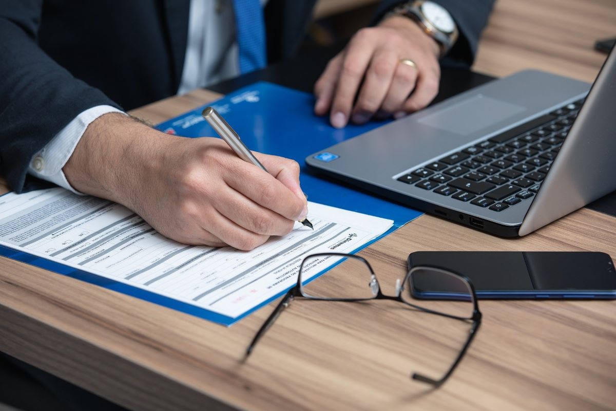 A man in a suit writing on a document next to his open laptop