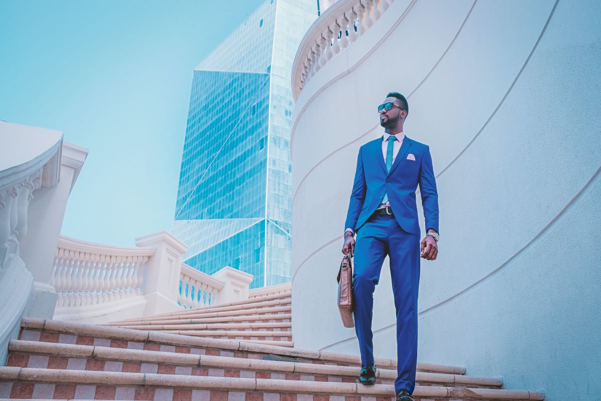 A well-dressed person in a suit strolls down the stairs and as they gaze up at the sky, with a glistening tall building as the backdrop.