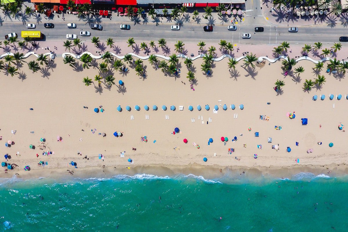 A drone shot of a Florida beach lined with palm trees and blue umbrellas, with people swimming in the water and relaxing on the sand