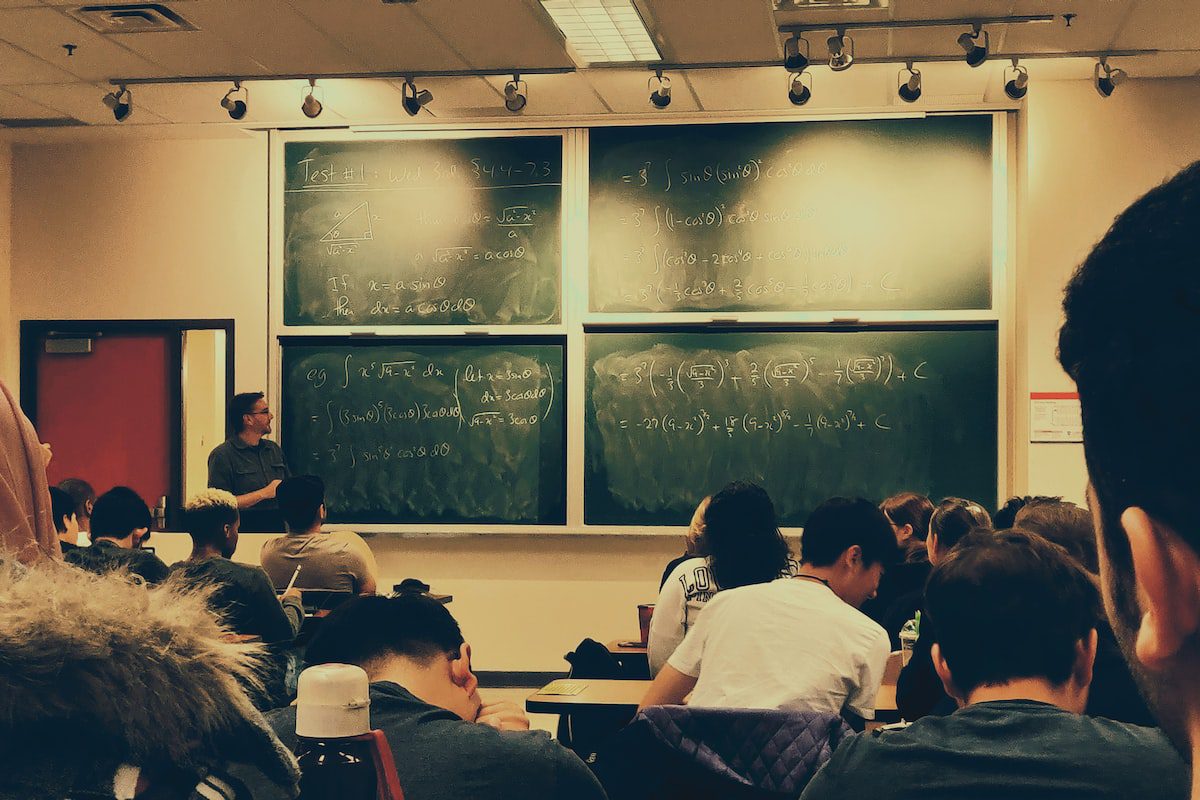 A college classroom full of students facing green chalkboards covered in mathematical equations as a professor lectures.