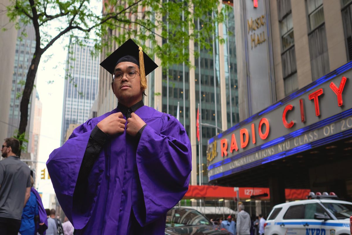 An NYU student in a purple graduation cap and gown standing in front of Radio City Music Hall in NYC.