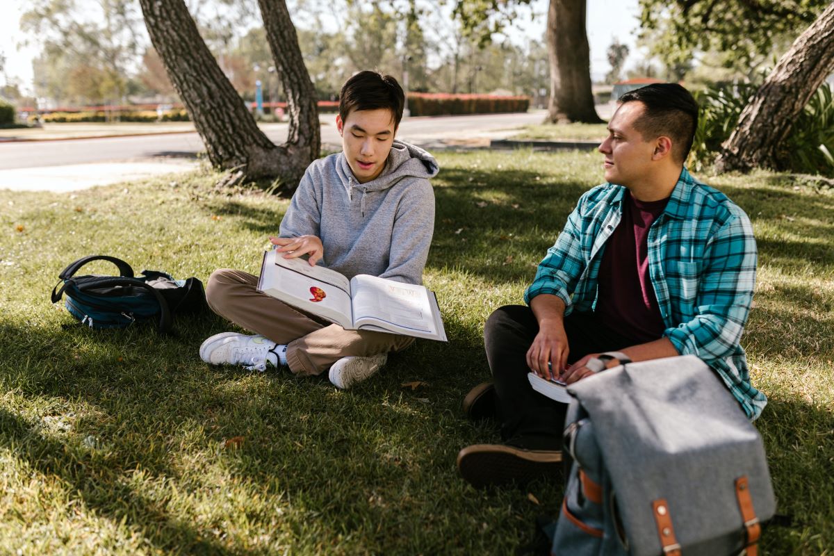 Two students sitting on a grass lawn looking at a textbook.