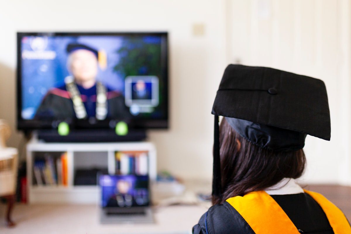 A student wearing a cap and gown attending an online graduation ceremony, their back is to the camera as they watch television.