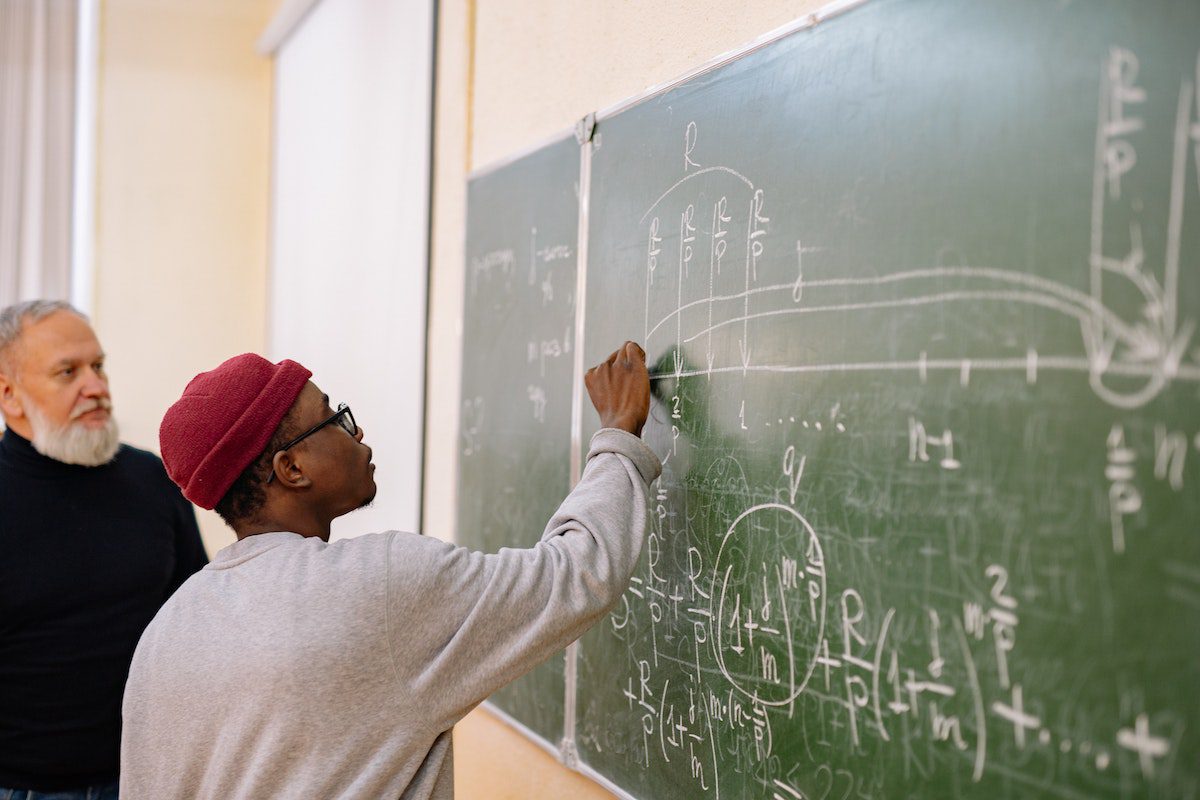 A student solving an equation on a blackboard while a professor watches.