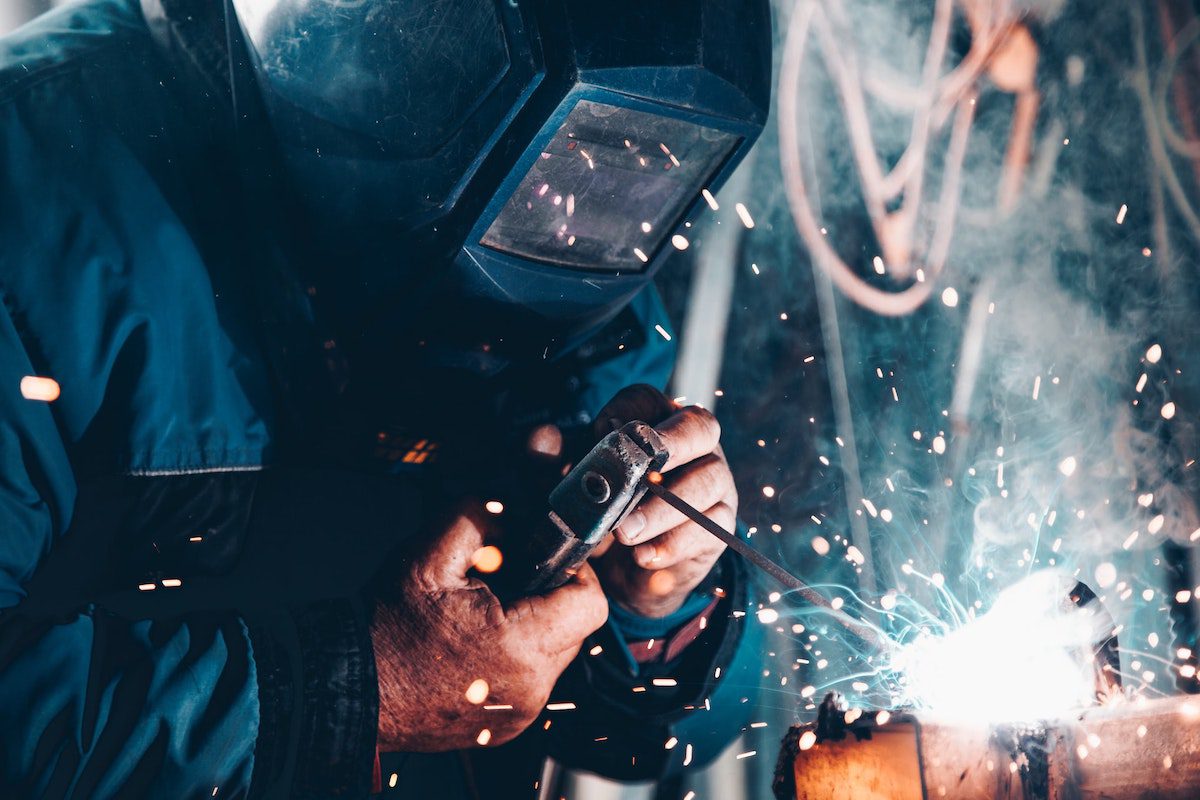 An engineer welding metal while wearing a blue jumpsuit and welding helmet.  